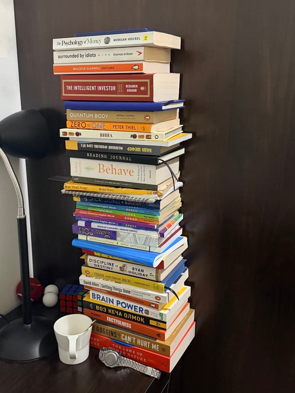 A waist-high stack of around thirty books on a desk beside a lamp — business, psychology and language titles in English, Russian and Uzbek
